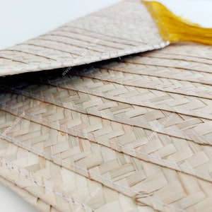 Op de afbeelding: Een gevlochten lichtbeige strooien clutch met een omgevouwen envelopklep en een felgeel kwastje. De buitenkant is gemaakt van natuurlijke vezels met zichtbare witte stiksels langs de horizontale gevlochten panelen.