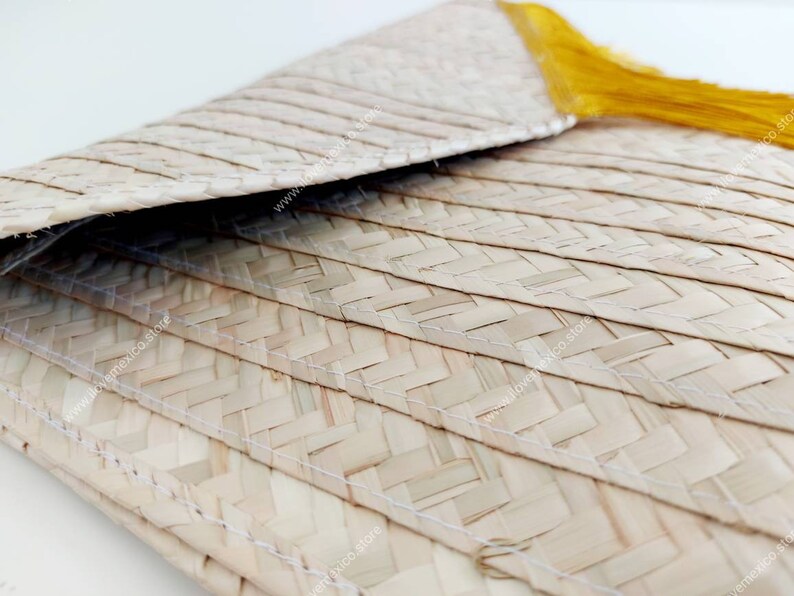 Op de afbeelding: Een gevlochten lichtbeige strooien clutch met een omgevouwen envelopklep en een felgeel kwastje. De buitenkant is gemaakt van natuurlijke vezels met zichtbare witte stiksels langs de horizontale gevlochten panelen.