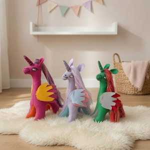 Puede incluir: Tres juguetes de unicornio de peluche hechos de fieltro, cada uno con un cuerno, alas y una larga melena. El unicornio de la izquierda es magenta con alas amarillas, el del centro es violeta claro con alas azul claro y el de la derecha es verde con alas grises.