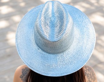 Puede incluir: Un sombrero fedora de color azul claro con una copa estructurada y un ala ancha y plana. El sombrero presenta una textura tejida en toda su superficie, creando un patrón consistente. El diseño incluye una parte superior pellizcada.