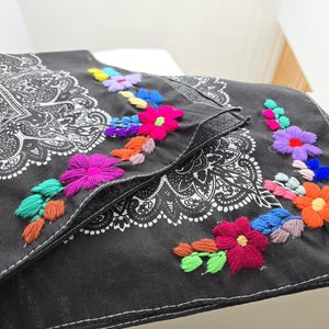 May include: A black fabric accessory featuring a white paisley print and vibrant floral embroidery. The embroidery includes multi-colored flowers in shades of pink, orange, blue, purple, and green, stitched along the edges of the fabric.