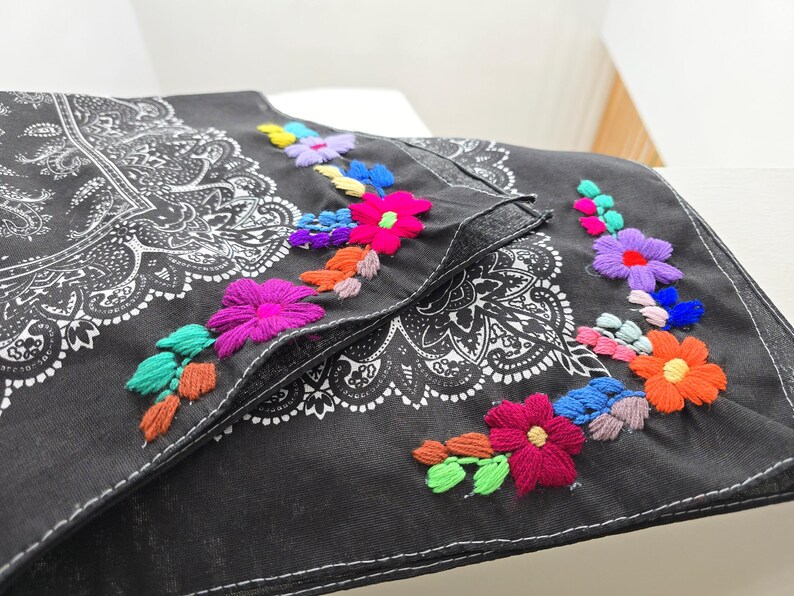 May include: A black fabric accessory featuring a white paisley print and vibrant floral embroidery. The embroidery includes multi-colored flowers in shades of pink, orange, blue, purple, and green, stitched along the edges of the fabric.