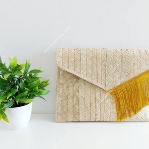 Op de afbeelding: Een rechthoekige clutch in envelopstijl, gevlochten van lichtbruine natuurlijke vezels in een visgraatpatroon. De flap is voorzien van een lange, levendige mosterdgele franje langs de diagonale rand van de sluiting voor een textuur.