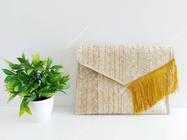 Op de afbeelding: Een rechthoekige clutch in envelopstijl, gevlochten van lichtbruine natuurlijke vezels in een visgraatpatroon. De flap is voorzien van een lange, levendige mosterdgele franje langs de diagonale rand van de sluiting voor een textuur.