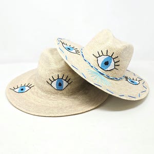 May include: Two straw hats with blue evil eye embroidery. The hats are beige and have a wide brim. The evil eyes are embroidered with black eyelashes and a blue pupil.