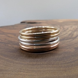 May include: A stack of six thin gold, rose gold, and silver rings on a wooden surface.