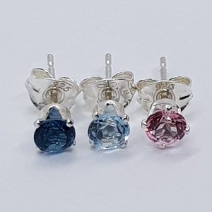 May include: Three pairs of sterling silver stud earrings with different colored gemstones. The earrings on the left have a dark blue gemstone, the middle earrings have a light blue gemstone, and the earrings on the right have a pink gemstone.