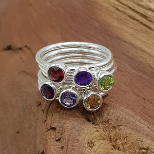 May include: A set of six silver stacking rings with colourful gemstones. The rings feature a red, purple, green, and yellow gemstone.