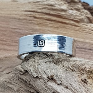 May include: A silver ring with a textured surface and a small square design on the top. The ring is sitting on a piece of wood.
