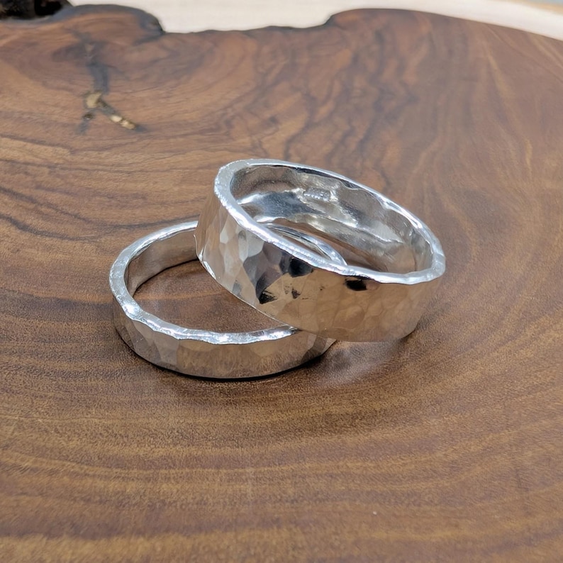 Pu&ograve; includere: Due anelli d'argento con una texture martellata sono impilati su una superficie di legno. Gli anelli hanno un semplice design a fascia e una superficie lucida e riflettente. Gli anelli sono probabilmente in argento sterling.