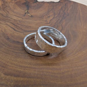 Pu&ograve; includere: Due anelli d'argento con una texture martellata sono esposti su una superficie di legno. Gli anelli sono rotondi con una superficie leggermente irregolare, che riflette la luce. Lo sfondo &egrave; una calda venatura del legno marrone scuro.
