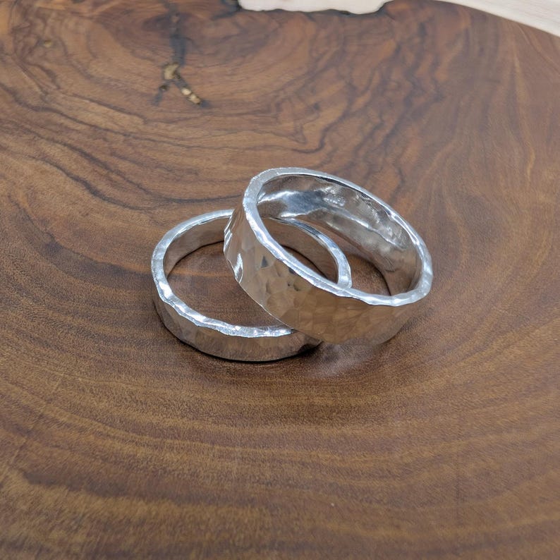 Pu&ograve; includere: Due anelli d'argento con una texture martellata sono esposti su una superficie di legno. Gli anelli sono rotondi con una superficie leggermente irregolare, che riflette la luce. Lo sfondo &egrave; una calda venatura del legno marrone scuro.