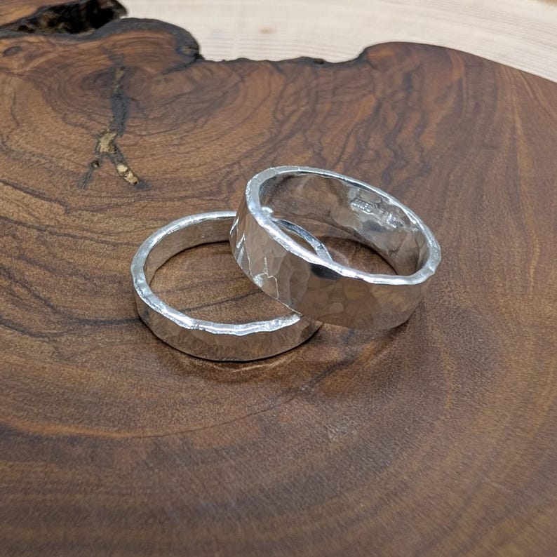 Pu&ograve; includere: Due anelli d'argento con una texture martellata sono mostrati su una superficie di legno. Gli anelli hanno un semplice design a fascia e una superficie leggermente irregolare, che riflette la luce. Gli anelli sono probabilmente per un matrimonio o un fidanzamento.
