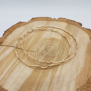 May include: Two gold-toned bangle bracelets with a wavy, textured design. The bracelets are stacked on top of each other on a wooden surface.