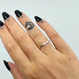 May include: A thin silver band ring with a hammered texture. The ring is worn on the ring finger of a hand with black nail polish.