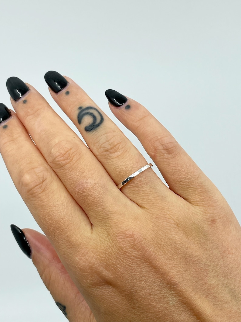May include: A thin silver band ring with a hammered texture. The ring is worn on the ring finger of a hand with black nail polish.