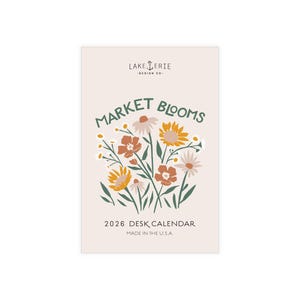 May include: A desk calendar with a cream background featuring a floral illustration in orange, yellow, and white. The text "Market Blooms" is above the flowers, and "2026 Desk Calendar" is below. The words "Made in the U.S.A." are at the bottom.