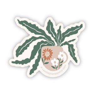 May include: A sticker featuring a green leafy plant growing out of a beige mug with a floral pattern. The mug has a white daisy, a large orange flower, and a green stem with leaves. The text "LAKEERIEDESIGNCO.COM" is at the bottom of the mug.