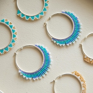 May include: Eight silver hoop earrings decorated with colorful seed bead patterns in turquoise, blue, gold, orange, and black. The earrings feature geometric designs and are arranged in a grid. The text Flores Clothing Boutique is visible.
