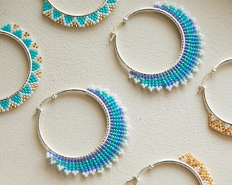 Puede incluir: Ocho pendientes de aro de plata decorados con patrones de cuentas de colores en turquesa, azul, dorado, naranja y negro. Los pendientes presentan diseños geométricos dispuestos en cuadrícula. El texto Flores Clothing Boutique es visible.