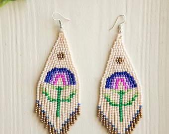 Puede incluir: Un par de pendientes colgantes hechos de pequeñas cuentas sobre una base de color crema. El diseño presenta un motivo floral central con cuentas azules, rosas y verdes, acentuado por una cuenta de bronce arriba y flecos de cuentas de bronce.