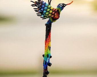 Può includere: Un ornamento sospeso a forma di colibrì, realizzato con piccole perline multicolori in un motivo arcobaleno. L'uccello presenta una lunga coda fluida composta da fili di perline con accenti neri, sospeso tramite un occhiello di perline nere.