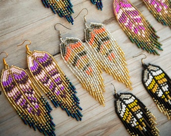 May include: Five pairs of long, dangling feather-shaped earrings crafted from small glass beads. The designs feature various color combinations including turquoise and gold, magenta and green, purple and gold, orange and silver, and black and yellow.