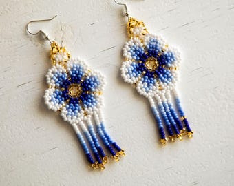 May include: A pair of dangle earrings crafted from small glass beads in a floral pattern. The petals feature a gradient of white, light blue, and dark blue beads, with a gold bead center. Each earring ends in a fringe of blue beads.