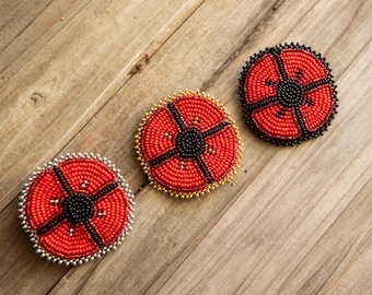 Peut inclure: Trois broches circulaires perlées représentant un motif de coquelicot rouge vif avec un centre noir. Chaque broche est ornée de petites perles, avec une bordure argentée à gauche, dorée au milieu et noire à droite.