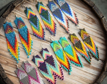 May include: A collection of six pairs of long, dangling seed bead earrings featuring intricate geometric chevron patterns in vibrant color combinations including blue, yellow, orange, pink, turquoise, black, and white, all with silver-toned metal hooks.