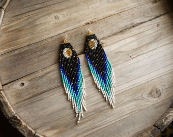 May include: A pair of long, dangling beaded earrings featuring a gradient design that transitions from black at the top to blue, turquoise, and white at the bottom. Each earring displays a circular gold and white moon motif near the top.
