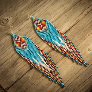 May include: A pair of long, dangling earrings crafted from small turquoise, white, red, orange, and yellow seed beads. The earrings feature a geometric diamond pattern at the top, transitioning into long, multi-colored beaded fringe strands.