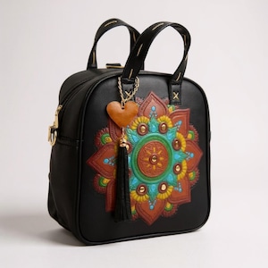 May include: A black handbag with two top handles and a zip closure. The front features a large, embossed, multi-coloured mandala design in shades of brown, yellow, and turquoise. A gold-tone chain holds a brown heart charm and a black tassel.