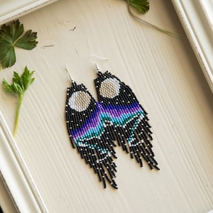 Puede incluir: Pendientes hechos a mano con cuentas con un diseño celestial. Los pendientes presentan un fondo negro con una luna creciente y un degradado de morado, azul y turquesa. La borla es negra con cuentas plateadas.