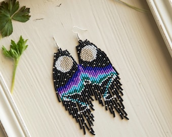 Puede incluir: Pendientes hechos a mano con cuentas con un diseño celestial. Los pendientes presentan un fondo negro con una luna creciente y un degradado de morado, azul y turquesa. La borla es negra con cuentas plateadas.