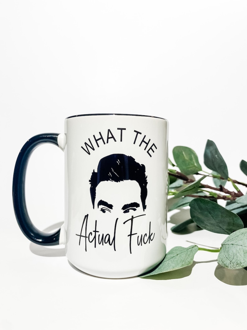 Puede incluir: Taza de cer&aacute;mica blanca con asa y borde azul oscuro. La taza presenta una ilustraci&oacute;n en blanco y negro de una cara y la frase "WHAT THE Actual Fuck" en letras negras.