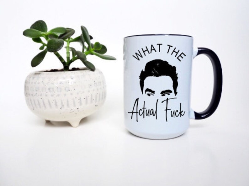 Puede incluir: Taza de cer&aacute;mica blanca con asa y borde negros, con la frase "WHAT THE Actual Fuck" y una silueta negra de una cara. A la izquierda, una peque&ntilde;a planta suculenta en maceta.