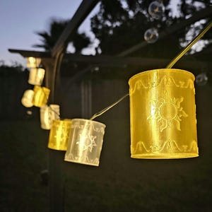 May include: A string of decorative lights featuring cylindrical lanterns with a sunburst design. The lanterns are illuminated in shades of yellow and white, strung along a thin wire, creating a warm, inviting glow. The lights are suitable for indoor or outdoor use.