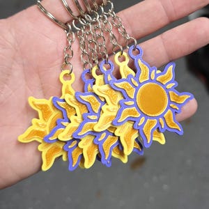 May include: A collection of keychains featuring a sun design. Each keychain has a yellow sun with a purple outline and a gold center. The keychains are attached to silver metal key rings and chains.