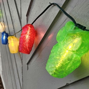 Puede incluir: Una cadena de luces con forma de búho de varios colores, incluyendo verde, rojo, amarillo, azul y blanco. Las luces cuelgan de una superficie de madera gris. Las luces están encendidas y parecen ser de plástico.