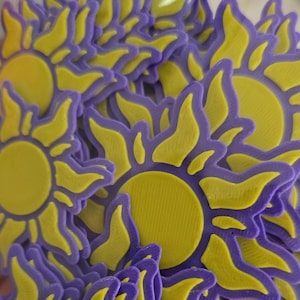 May include: A collection of yellow and purple sun-shaped objects. Each sun has a yellow center and purple rays. The objects appear to be 3D printed or molded, with a slightly textured surface. The overall theme suggests a craft or decorative item.