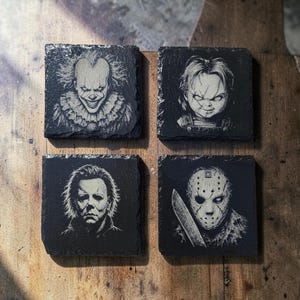 May include: A set of four square black slate coasters featuring laser-engraved portraits of horror movie characters. The designs include Pennywise the clown, Chucky the doll, Michael Myers, and Jason Voorhees with a machete, all in high-contrast white.
