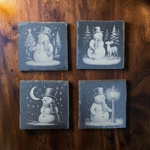 May include: A set of four square slate coasters featuring white engraved winter scenes. Each coaster displays a snowman wearing a top hat and scarf, depicted with a Christmas tree, a deer with a lantern, a crescent moon, or a street lamp.