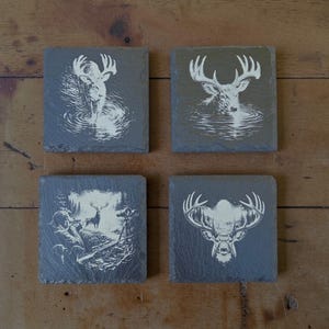 May include: A set of four square dark gray slate coasters, each featuring a white engraved illustration of a deer. The designs include a deer in water, a deer head with antlers, and a hunter observing a deer in a forest setting.
