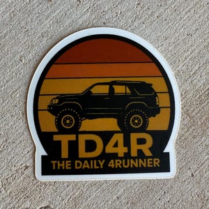 May include: A die-cut sticker with a black SUV silhouette set against an orange and yellow sunset gradient. The text "TD4R THE DAILY 4RUNNER" is in white on a black banner, ideal for car enthusiasts.