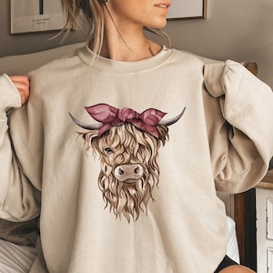 May include: Beige sweatshirt featuring a watercolor illustration of a Highland cow with a pink bow. The cow has long, shaggy brown fur and curved horns. The sweatshirt has long sleeves and a crew neck.