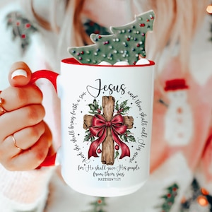 May include: White ceramic mug with a red interior and handle, featuring a wooden cross design with a red bow and the word "Jesus." The mug also includes text from Matthew 1:21 and is topped with a green Christmas tree cookie.