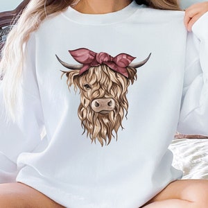 May include: White sweatshirt featuring a watercolor illustration of a Highland cow with a pink bandana. The cow has long, shaggy brown fur, dark horns, and a light brown face. The sweatshirt is a crew neck style.