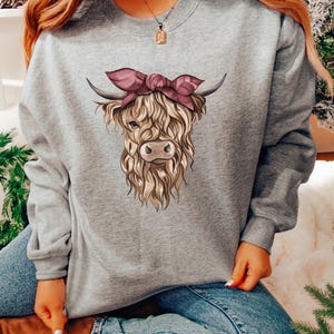 May include: A heather gray sweatshirt featuring a detailed illustration of a Highland cow. The cow has long, shaggy fur, horns, and a pink bow. The sweatshirt is a comfortable, casual style.