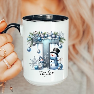 May include: White ceramic mug with a black interior and handle. The mug features a winter-themed design with a large letter "T" decorated with blue ornaments, snowflakes, and a snowman. The name "Taylor" is printed below the design.